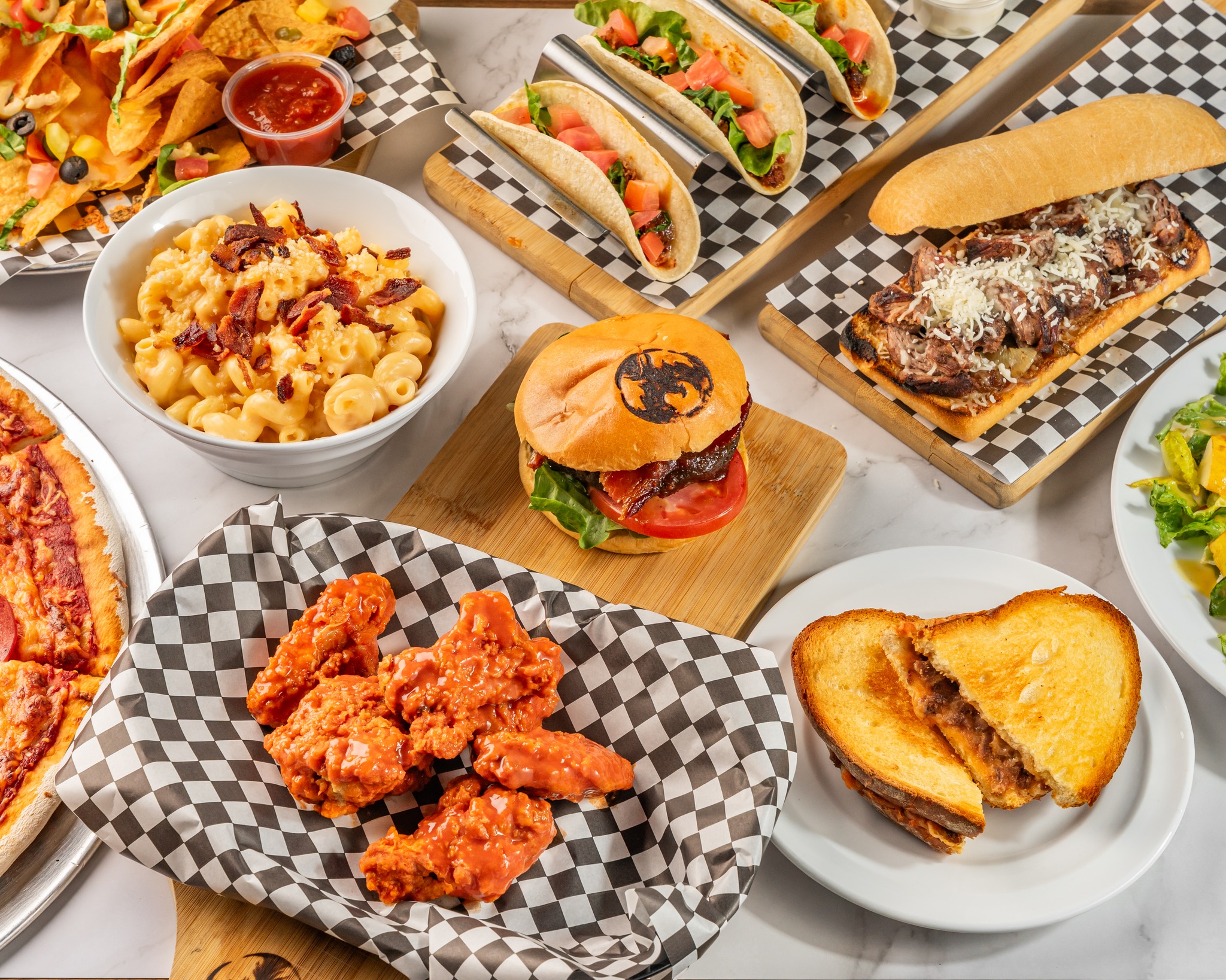 Spread of Ignition Eatery dishes including burgers, tacos, mac and cheese, wings and flatbread on checkered deli paper